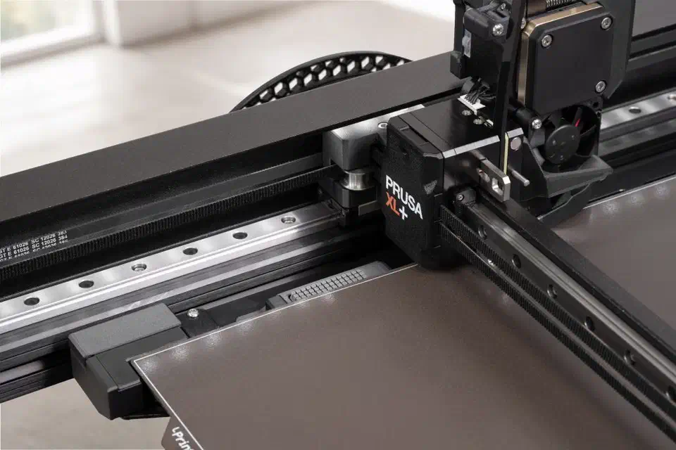 Close-up of the Prusa XL+ toolhead and belts with the eddy-current tool offset sensor strip set into the front edge of the bed