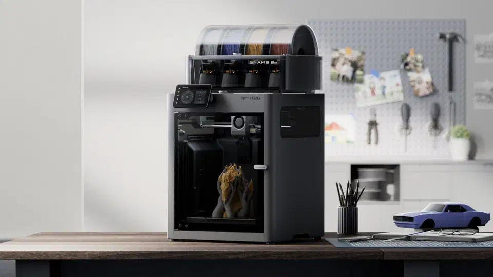 Bambu Lab X2D printer on a wooden desk with a four-spool AMS 2 Pro on top and a two-colour model printing inside the lit chamber