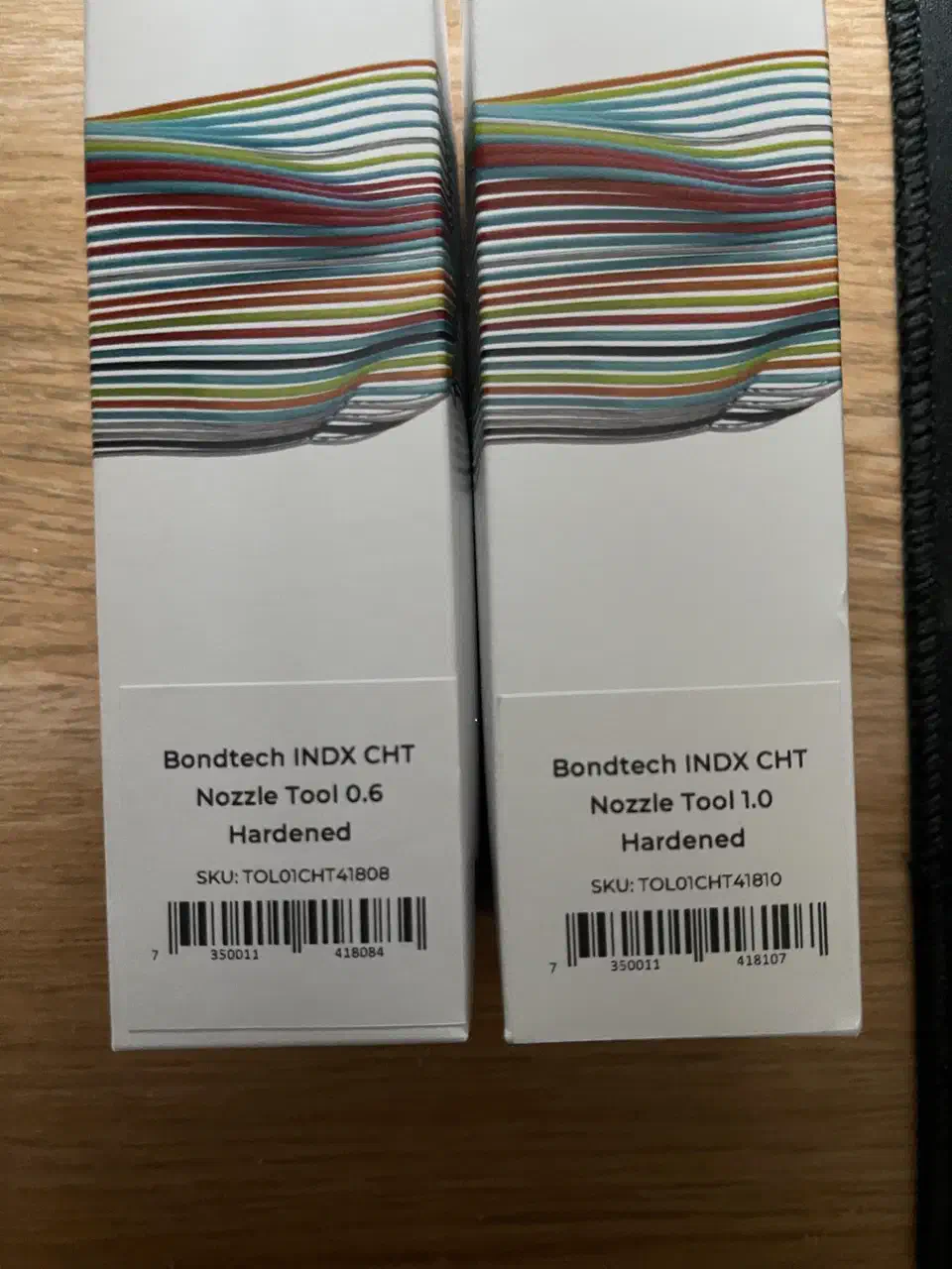 Two white Bondtech retail nozzle boxes on a wooden desk, labelled Bondtech INDX CHT Nozzle Tool 0.6 Hardened and 1.0 Hardened with SKU barcodes