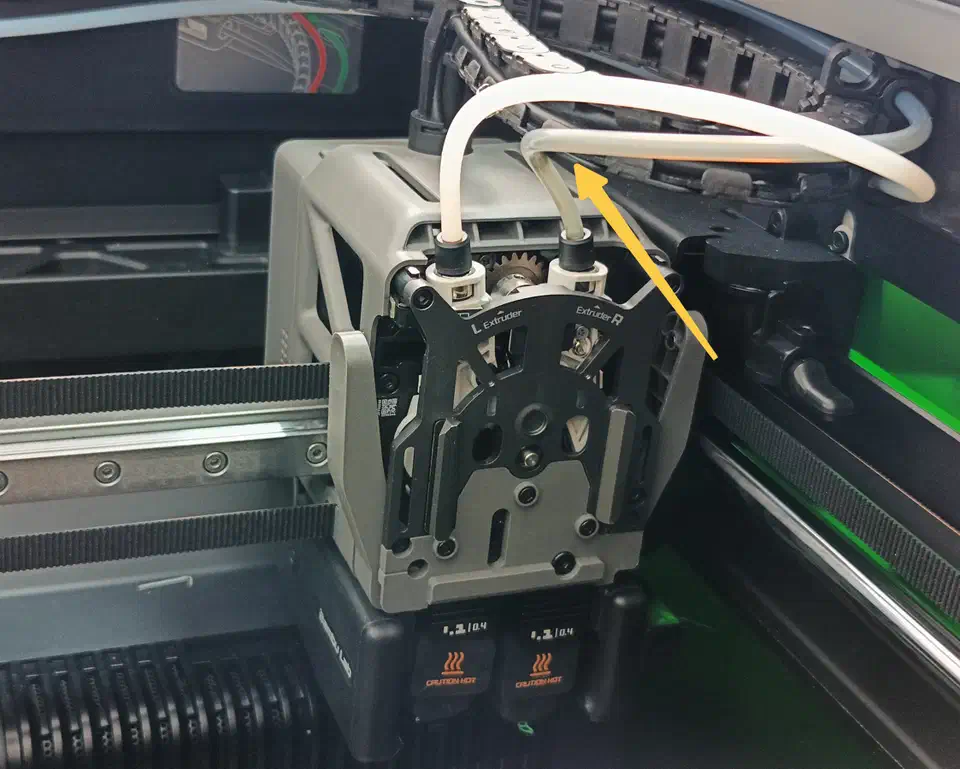 Close-up of the H2D dual extruder with the filament tube routed through the feed path, an arrow marking the securing point brittle filaments need