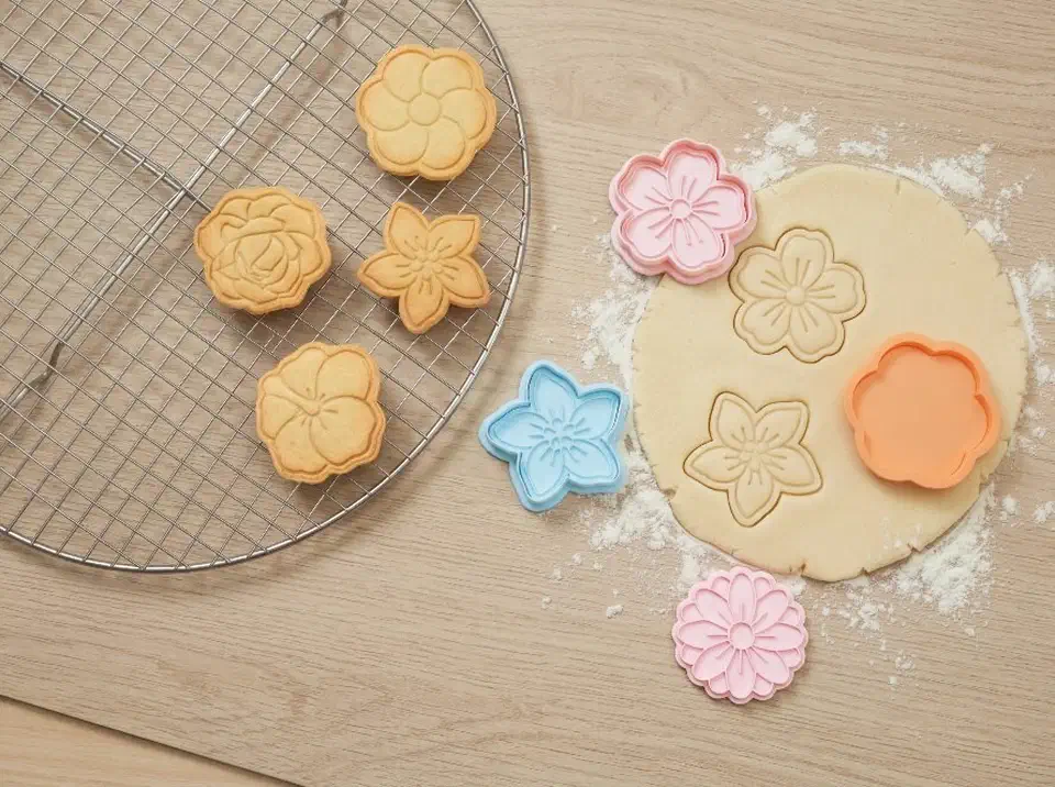 Pink, blue, and orange 3D-printed flower cookie cutters pressing shapes into dough beside baked cookies on a cooling rack