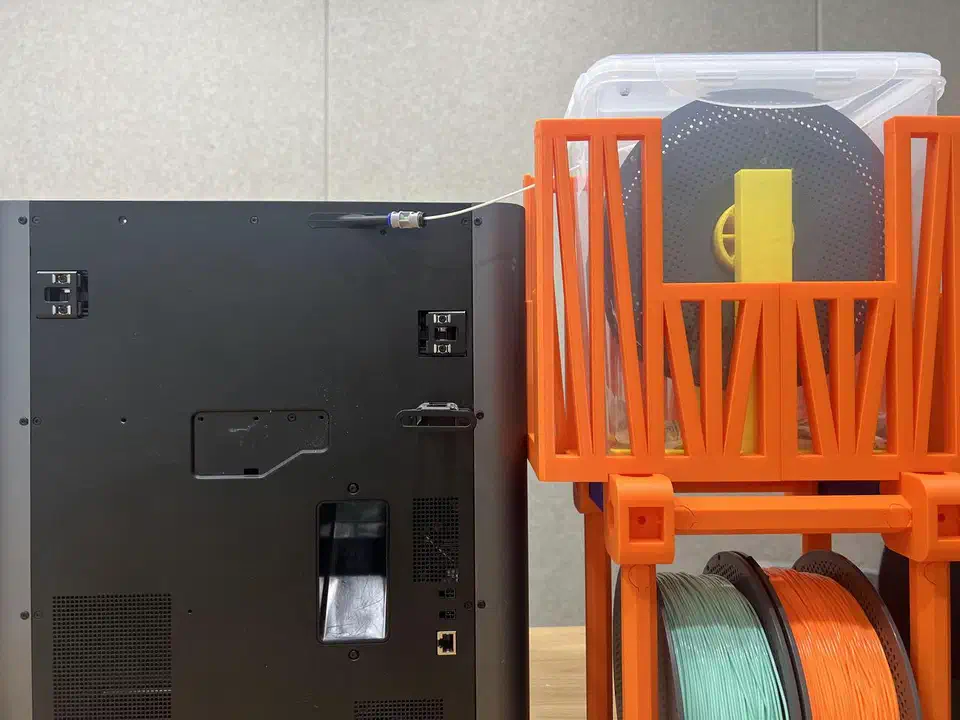 A carbon-fiber filament spool sealed inside a clear airtight dry box feeding an enclosed printer, with two more spools stored below