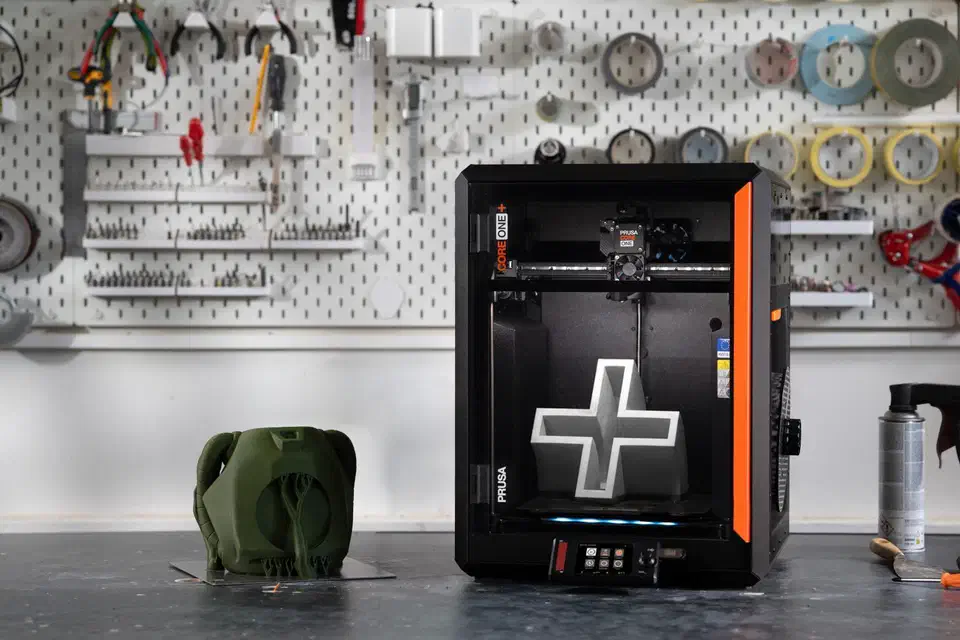 Black enclosed Prusa CORE One+ printer with orange front accent on a workshop bench, printing a large white cross, beside a green printed part and a pegboard of tools