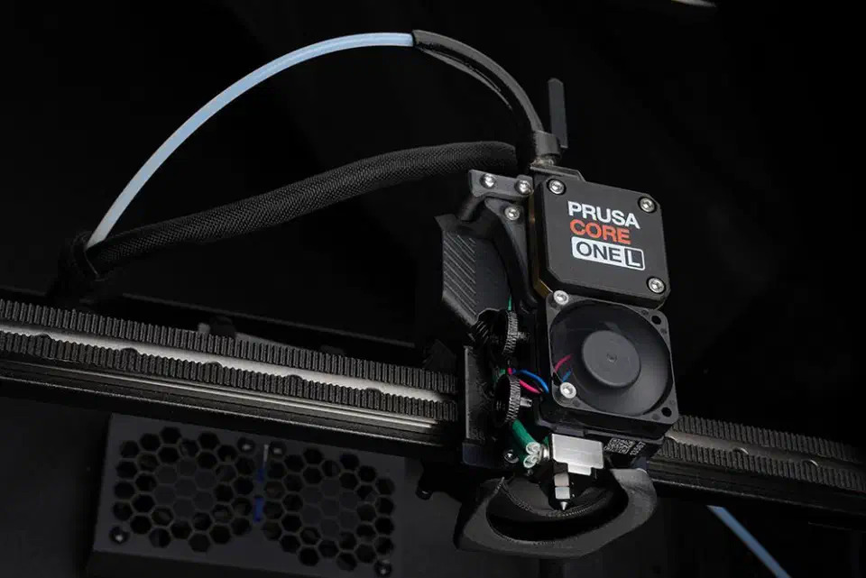 Prusa CORE ONE L toolhead close-up mounted on the gantry belt, with cooling fan, filament tube and nozzle visible inside the printer
