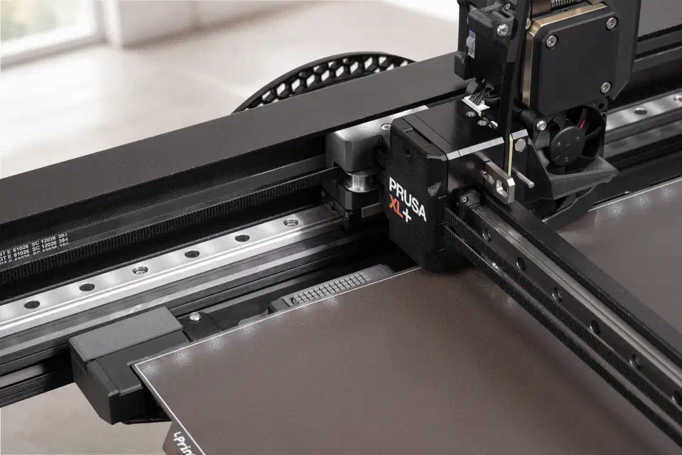 Prusa XL+ gantry close-up showing the black toolhead carriage, linear rails, drive belt and the wiper block beside the print sheet