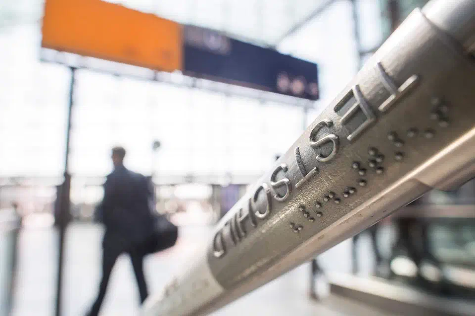 Metal train station handrail with embossed lettering and braille dots, a commuter walking in the blurred background