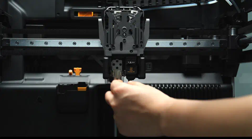 A hand pulling a hotend module out of the dual extruder toolhead inside an enclosed Bambu printer, with a caution hot label on the module