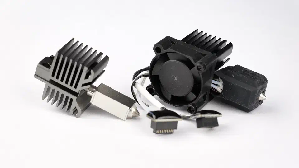 A bare nozzle and heatsink beside a full hotend assembly with its cooling fan, silicone sock and ribbon connectors laid out on a white surface
