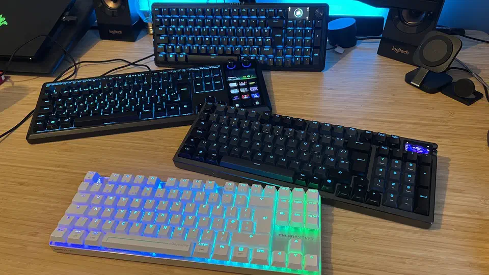Four Hall effect gaming keyboards laid out on a wooden desk, including the Asus ROG Azoth 96 HE with its ROG display, a Corsair Vanguard Pro 96, and a white Cherry XTRFY board