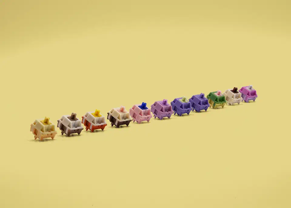 A row of HMX linear switches in many colours, each with a coloured housing and a cross-shaped stem, lined up on a yellow background