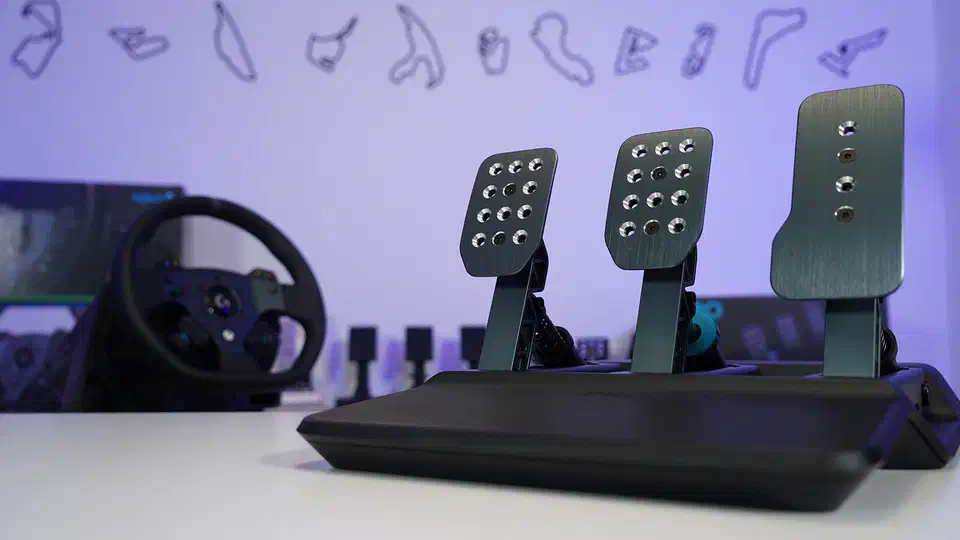 Logitech PRO Racing Pedals on a white desk, three tall brushed metal pedal faces on a black base with a racing wheel blurred behind them