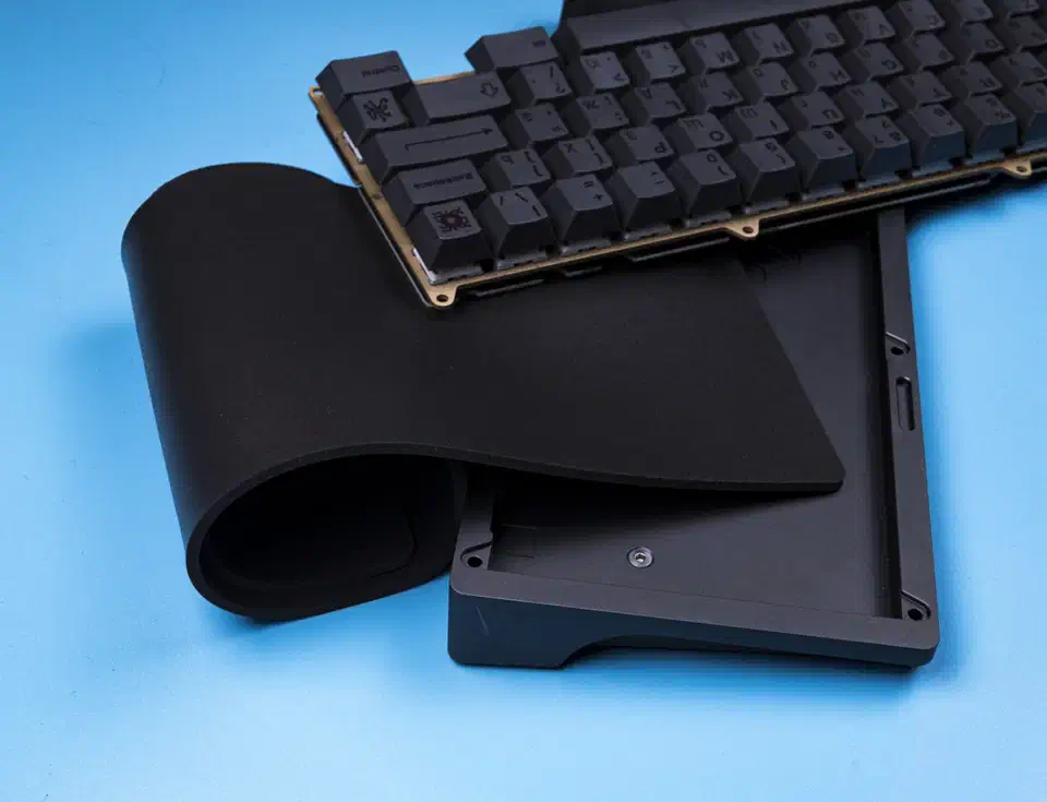 A black Poron case foam sheet curling out of an empty dark grey keyboard case, with the assembled plate, PCB and black keycaps resting beside it