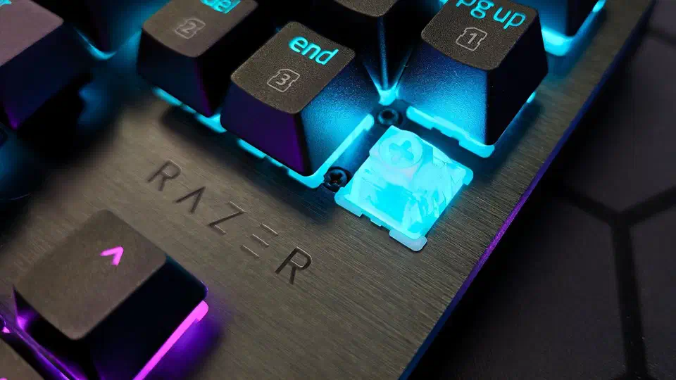 Close-up of a translucent Hall effect magnetic switch on the Huntsman V3 HE with its keycap removed, lit cyan, beside the brushed aluminium Razer logo plate