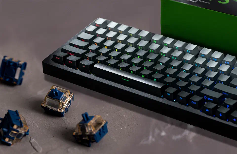 MonsGeek M1 V5 TMR keyboard with backlit keycaps beside four loose blue and gold Akko Astrolink magnetic switches on a grey surface