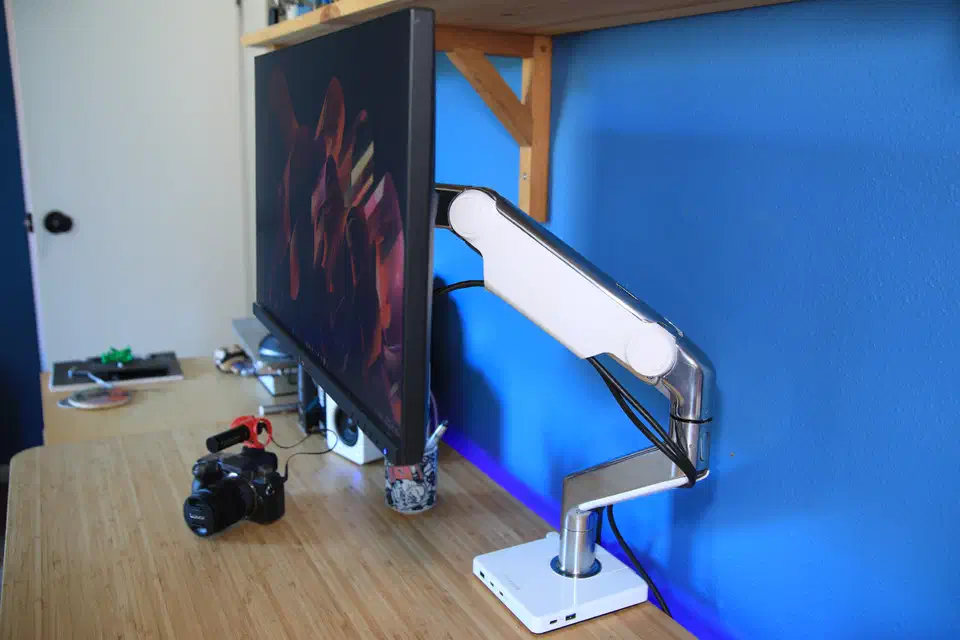 Silver Humanscale M2.1 heavy-duty monitor arm holding a display, cable run down the arm to a white USB dock at the desk clamp