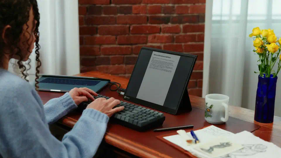Modos Flow standing on a wooden desk beside a mechanical keyboard, showing a page of dark text on its pale e-paper panel
