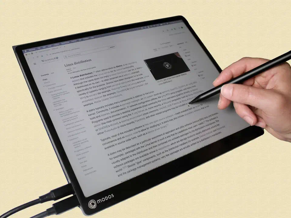 Hand holding a black stylus against the Modos Flow screen, which is showing a Wikipedia article in grey tones