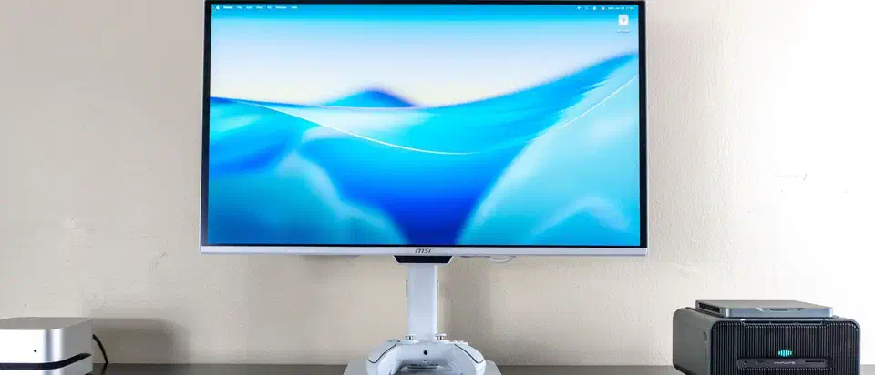 White MSI Pro Max 27-inch QD-OLED monitor on a desk between two small computers, showing a blue macOS desktop
