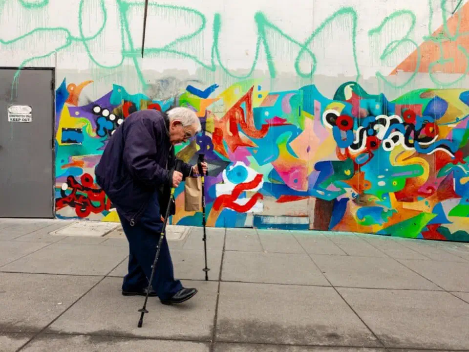 Street scene shot on the Galaxy S26 Ultra main camera, a man with walking sticks passing a painted wall mural