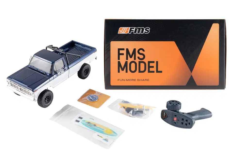 FMS FCX18 II F-100 truck beside its orange FMS box, FlySky transmitter, decal sheet with surfboard, and bagged accessories