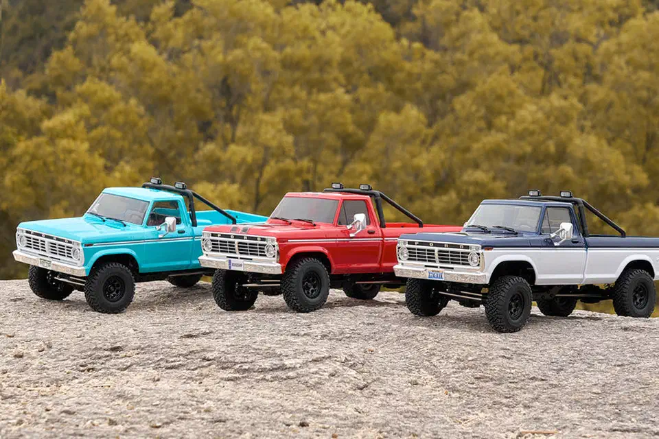 Turquoise, red and navy-and-white FMS FCX18 II Ford F-100 crawlers parked in a row on a rock slab in front of trees