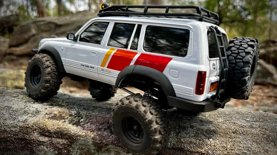 Rear three-quarter view of the FCX18 LC80 crawler on rock, showing the tailgate-mounted spare tire, roof rack and knobby portal-axle tires