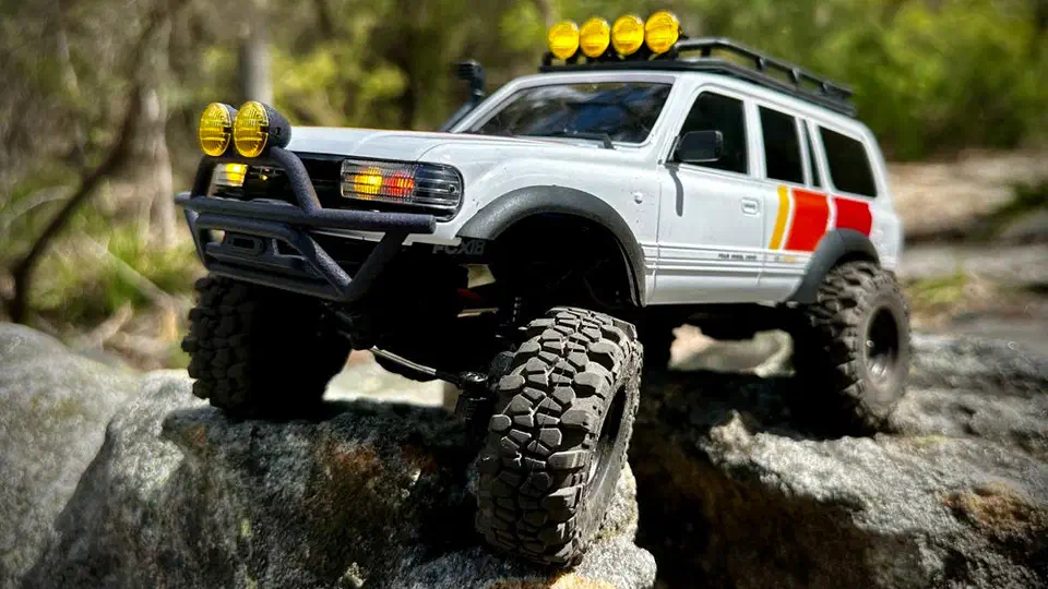 FMS FCX18 LC80 tri-color crawler perched on a granite boulder, front bumper light pods and roof light bar visible above the tri-color side stripes
