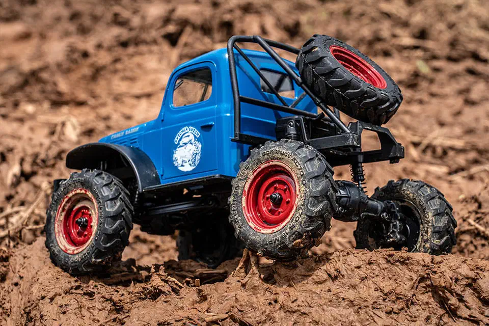 Rear three-quarter view of the blue FCX24 Power Wagon flexing on mud, showing the four-link suspension, coil shocks, and spare tire on the roll cage